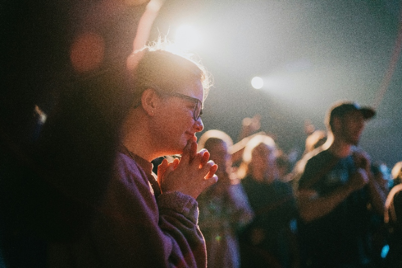 Young people engaged in worship in a concert style setting. One woman is in focus in the foreground; soft focus of 3 more people (2 women, i man) in the background