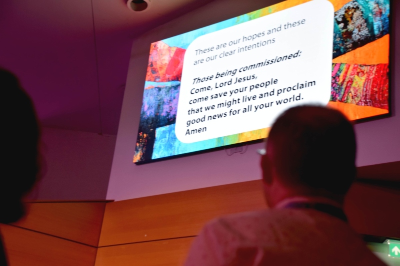 A photo from the back of the head of a newly commissioned male minister in front of a screen detailing one of the promises he is invited to make