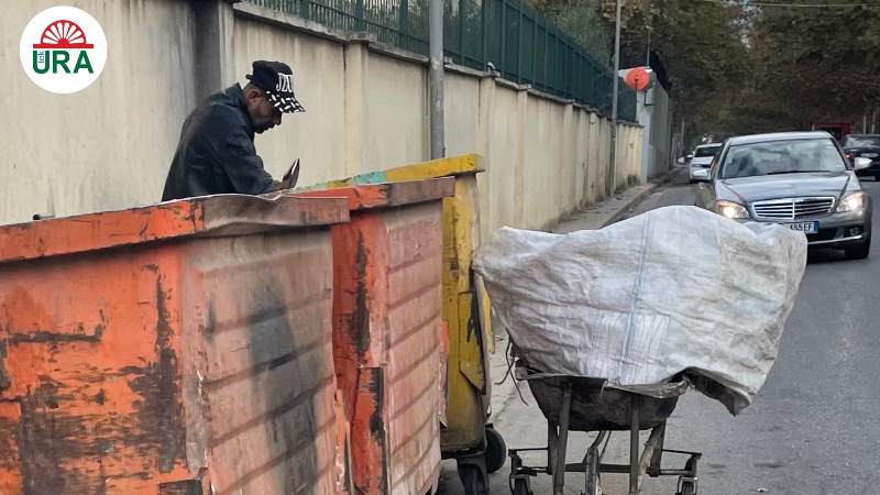 Albanian street, a man is rooting through a rubbish bin while cars drive past
