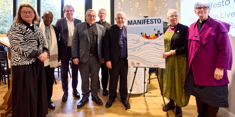 8 church leaders in Merseyside (5 men, 3 women) stand in a line next to a Faith in the City Manifesto poster