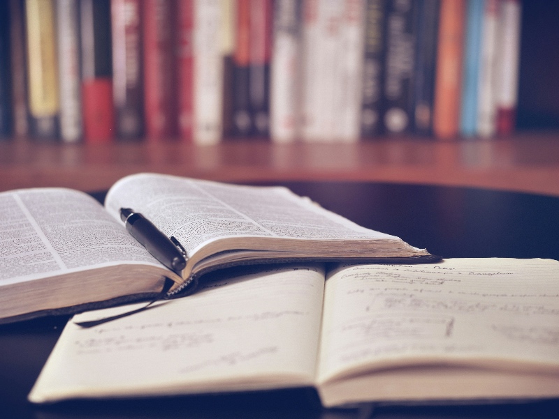 An open Bible with a fountain rests on an open notebook against a backdrop of a shelf of books