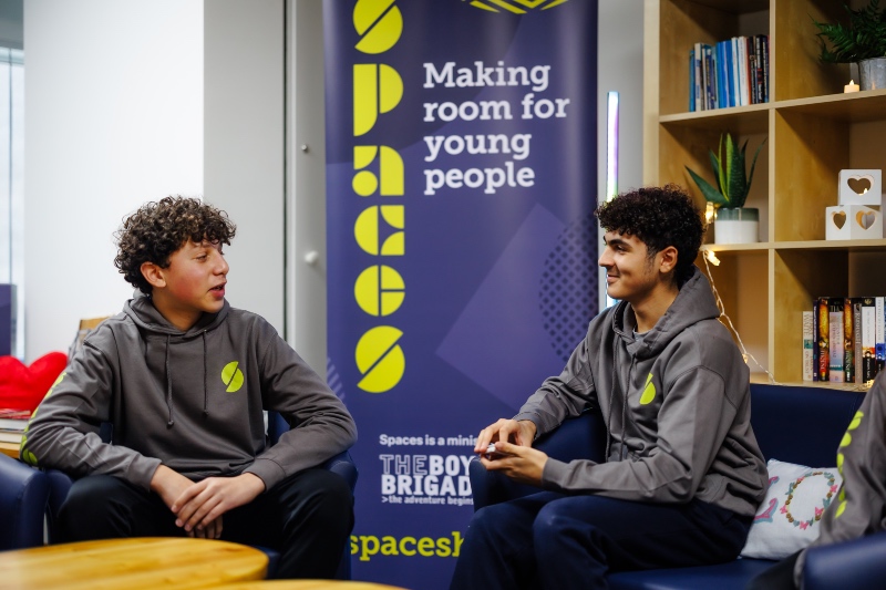 Two young people in matching grey hoodies sit and talk in a lounge area with a banner behind them that reads “Spaces - Making room for young people."