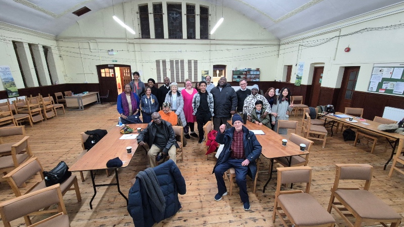 A group of people pose for a photo in a large church hall with wooden floors, surrounded by tables, chairs, and papers