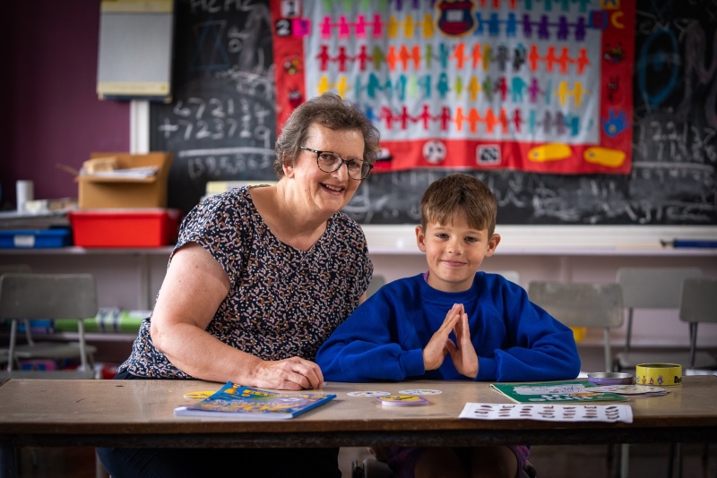 A smiling woman and young boy sit at a classroom table with educational materials. Blackboard and colourful wall decor are visible in the background