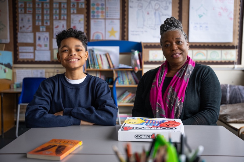 A boy and a woman sit at a table in a classroom. A book, a game of Connect 4, and a tub of felt tips are on the table
