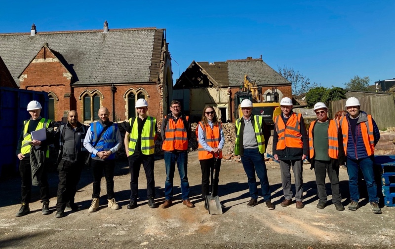 A group of ten people wearing hard hats and high-visibility vests stand in a row at a construction site. There's a historic brick building and clear blue sky in the background. The photo marks the beginning of a £3.4m building project of Christ Church New Southgate and Friern Barnet Baptist / United Reformed Church 