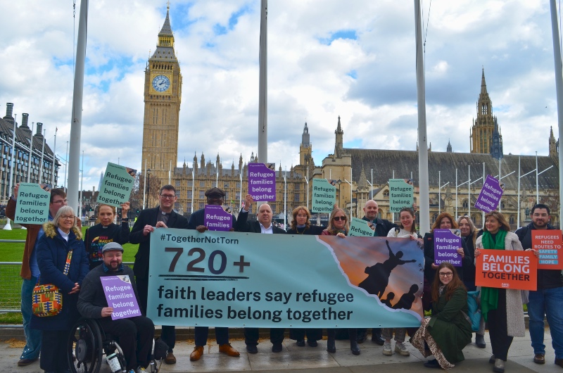 A diverse group holds a banner reading "720+ faith leaders say refugee families belong together" outside UK Parliament. Signs state "Refugee families belong together".  Big Ben is visible, under a cloudy sky.