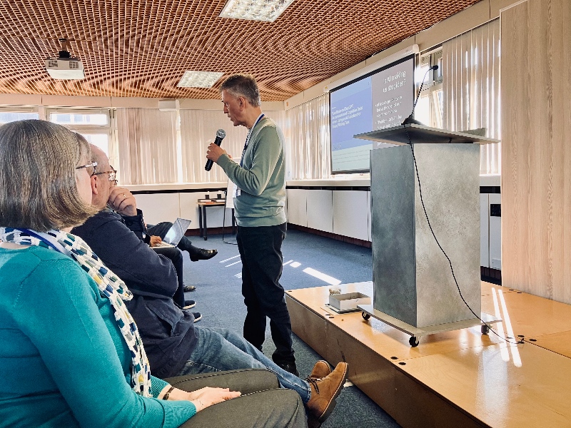 Chris Goswami speaking into a microphone beside a podium in a conference room, with an attentive seated audience and a projection screen in the background.