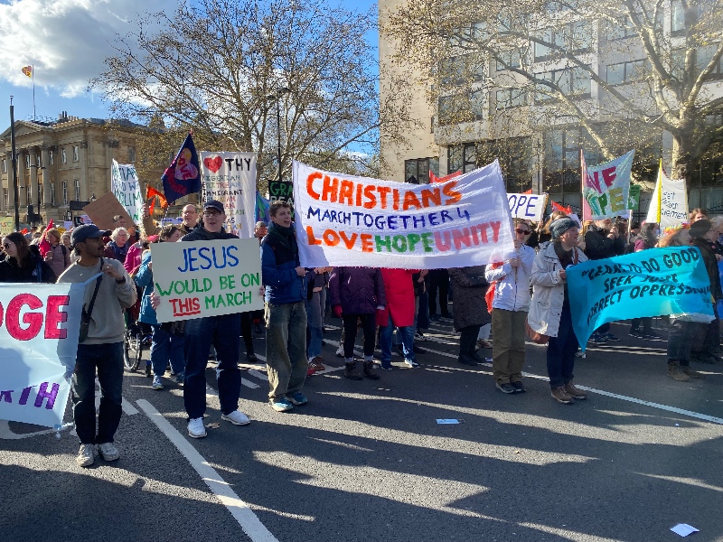 A diverse group of Christians march in a peaceful protest in London, holding banners with messages of love, hope, and unity.