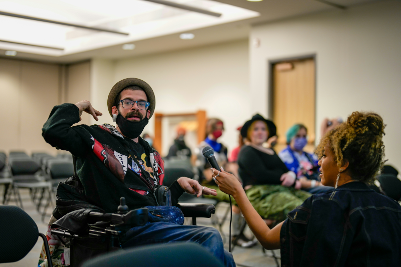 A man in a hat and glasses, seated in a wheelchair, speaks into a microphone held by a woman with curly hair. Others sit in rows of chairs