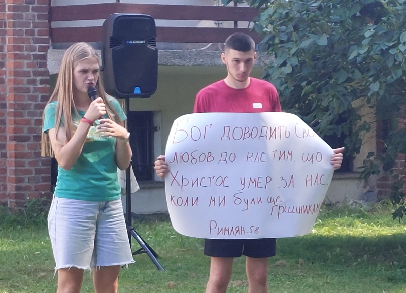 A woman in a green shirt speaks into a microphone outdoors. Next to her, a man holds a handwritten sign with text. They stand on grass near a brick wall.