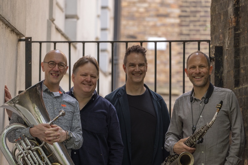 Four smiling men - the members of the Kairos Ensemble - pose with musical instruments, a euphonium and a saxophone, against a gated alleyway backdrop