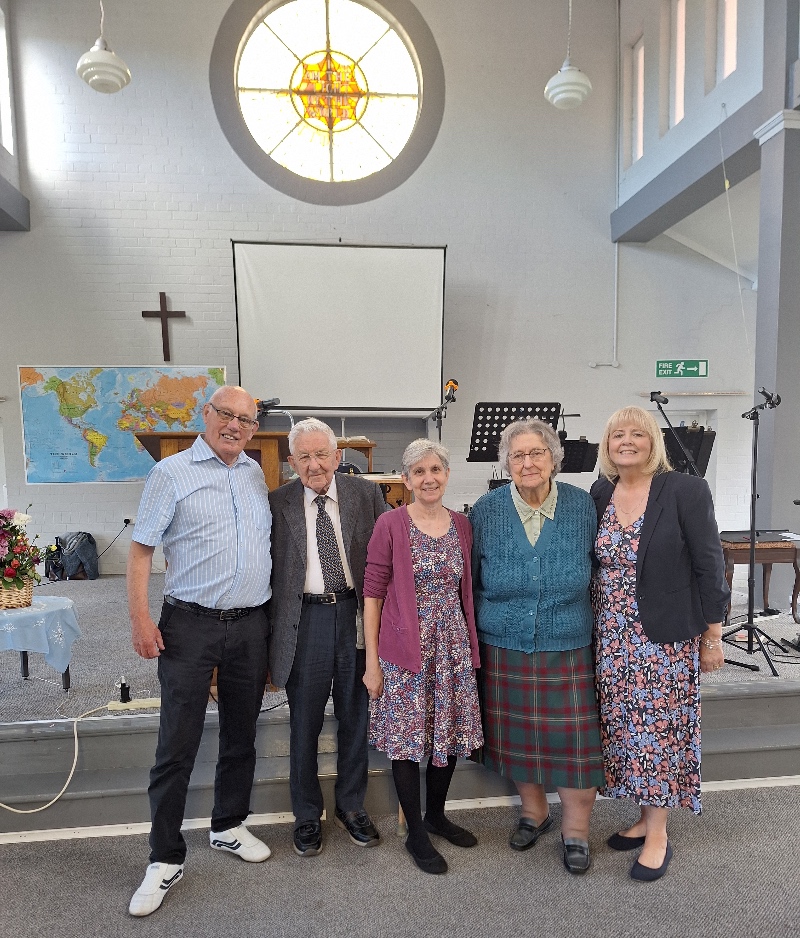A group of five people, smiling and standing inside a church with a cross, stained glass window, and world map.
