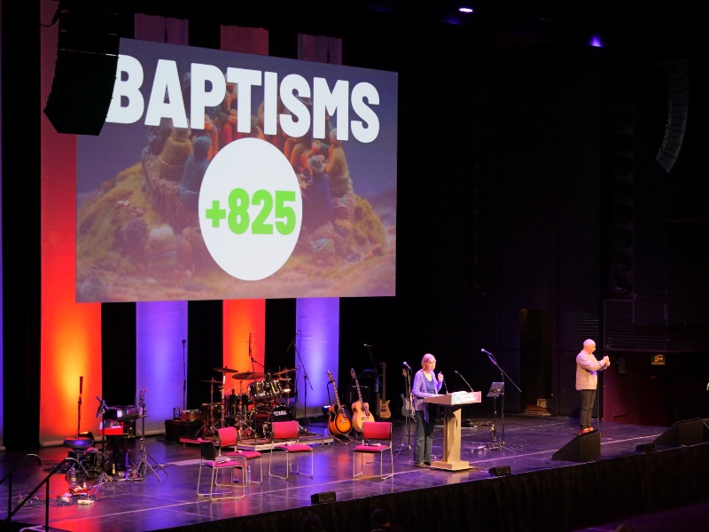 The Baptist Assembly stage with a large screen displaying "Baptisms +825." Lynn Green is speaking at a podium, with musical instruments in the background.