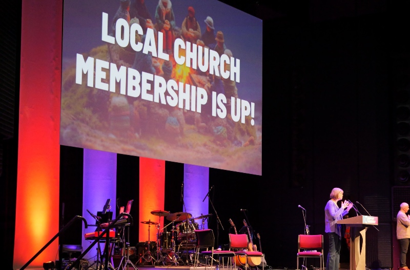 Lynn Green stands at a podium on the Baptist Assembly stage beside a large screen displaying "Local Church Membership is Up!" The stage is lit with vibrant orange and purple lights Musical instruments are visible in the background.