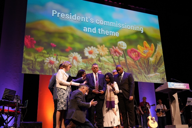 Alex Afriyie is prayed for by a group of people, under a colourful floral backdrop that says “President's commissioning and theme”