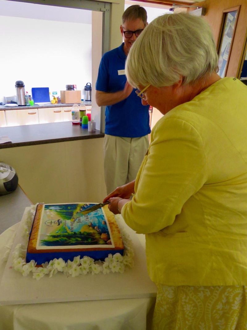 A woman in a yellow outfit (MP Charlotte Cane) cuts a decorated cake at a celebration, with a smiling man (minister Chris Johnson) in a blue shirt observing.