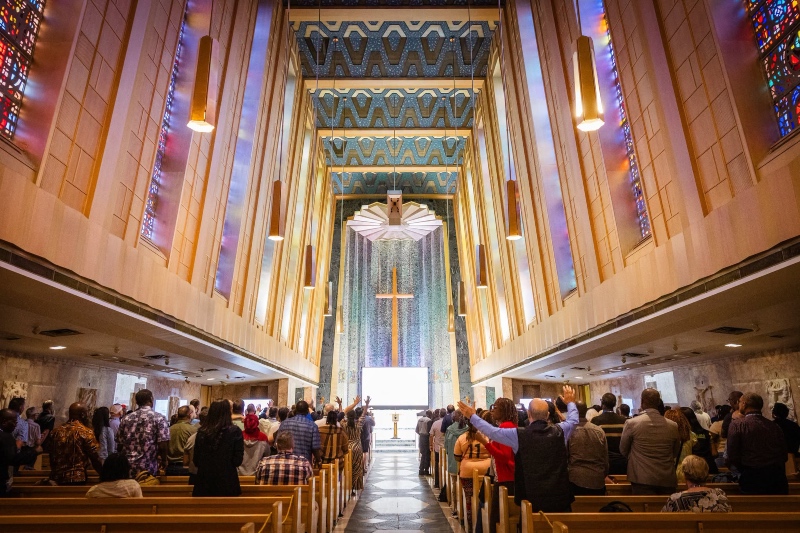 Sung worship at the BWA annual gathering - A view from the back of a modern cathedral with high, angled wooden walls, showing a congregation standing with raised hands in worship toward a central wooden cross.