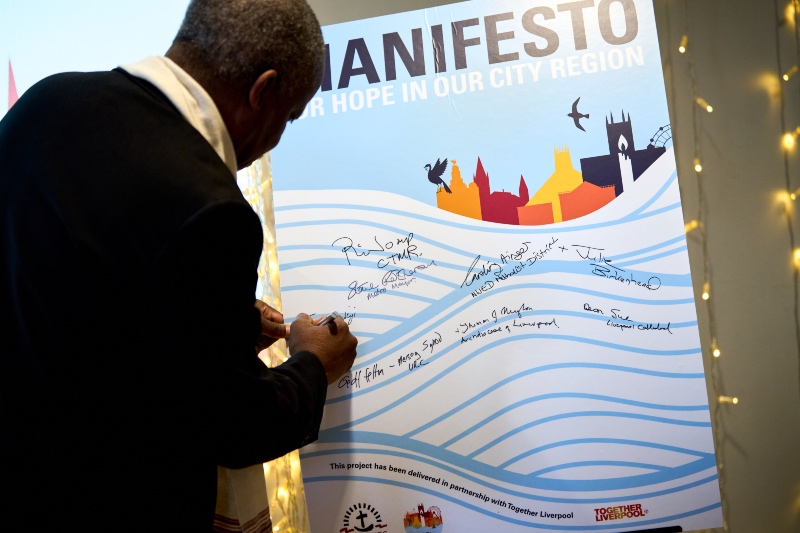 A close-up view of a man signing a large poster titled "MANIFESTO FOR HOPE IN OUR CITY REGION," which features city skyline graphics and multiple signatures.