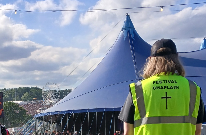 A person wearing a high-visibility vest labeled "FESTIVAL CHAPLAIN" looking toward a large blue festival tent with a Ferris wheel in the background.