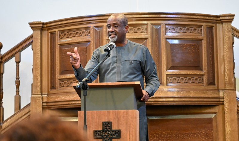 Karl Johnson speaking at a wooden lectern with a microphone, gesturing with his hand in front of an ornate wooden church pulpit.