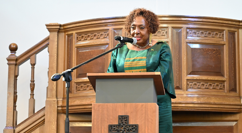 Olivia Grange, in a green dress, speaking at a lectern with a microphone in front of an ornate wooden church pulpit.