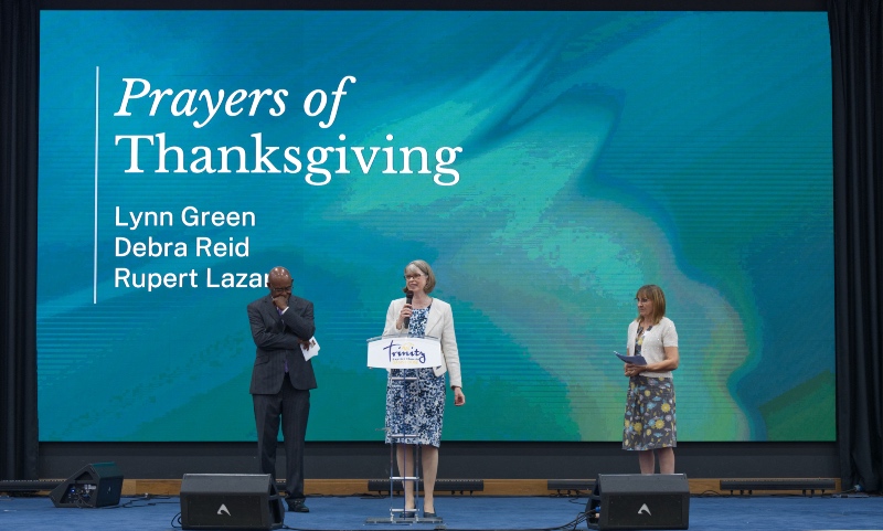 Three speakers standing on a stage in front of a large screen displaying "Prayers of Thanksgiving" and the names Lynn Green, Debra Reid, and Rupert Lazar.