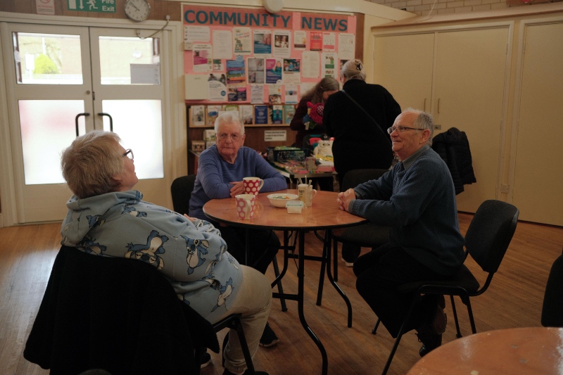 Three adults sitting around a round table chatting over hot drinks in a community hall